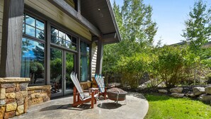 Une maison avec de grandes fenêtres, un mur en pierre et une terrasse dotée de deux fauteuils Adirondack et d’un foyer extérieur.