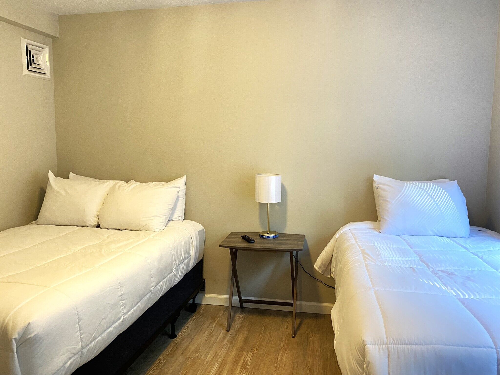 Una habitación de hotel con dos camas individuales, una mesita de noche con lámpara y un respiradero en la pared.