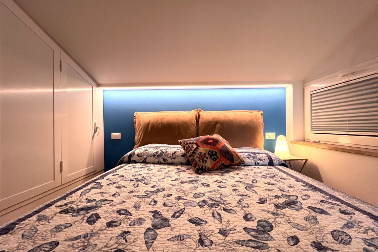 Een slaapkamer met een bed, kussens, een nachtkastje en een raam met jaloezieën.