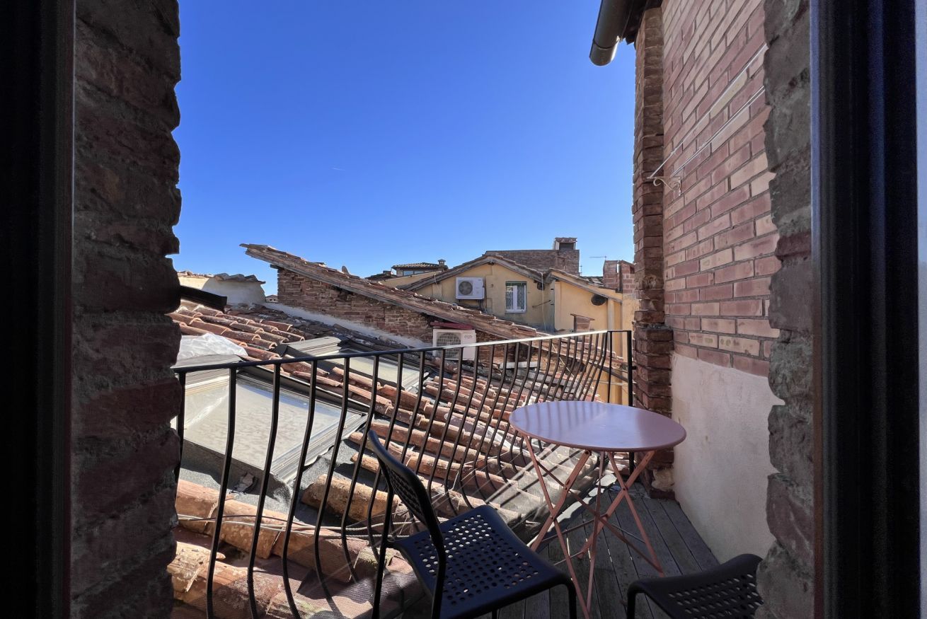 Un balcón con una pequeña mesa y sillas, con vista a un patio que tiene una pared de ladrillo y un edificio al fondo.