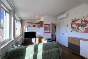 A well-lit room with a bed, a TV, and a kitchenette. There is a window providing natural light and a door leading to another room.