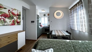 Une chambre à coucher de taille modeste, comprenant un lit, une table de chevet, un luminaire rond fixé au mur, et donnant sur une cuisine et un coin repas.