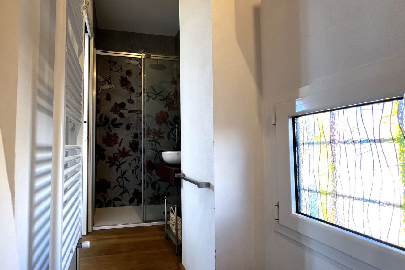 Een smalle gang met een houten vloer, een douchedeur van glas met bloemenbehang, en een raam met uitzicht op bomen.