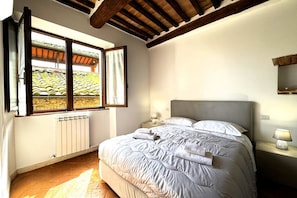 A bedroom with a large bed, a window with a view of a garden, a radiator, and wooden beams on the ceiling.