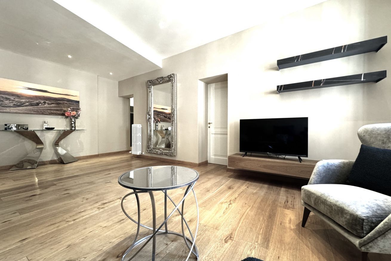 A modern living room with a round glass table, a large mirror, a flat-screen TV, and a comfortable armchair.