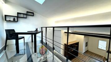 A modern apartment with a glass staircase, a dining area with a black chair, and a kitchen visible through the glass.