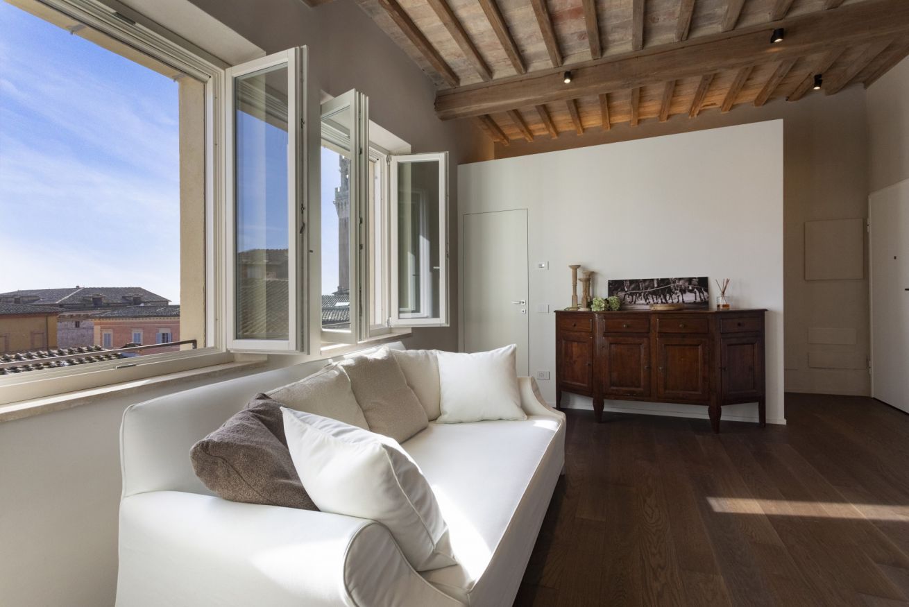 A modern living room with a white sofa, wooden ceiling beams, a large window with an open door, and a view of buildings outside.