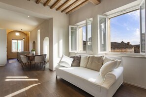 Una sala de estar moderna con un sofá blanco, una mesa de comedor y sillas. Se ve un ventanal con vistas a edificios.