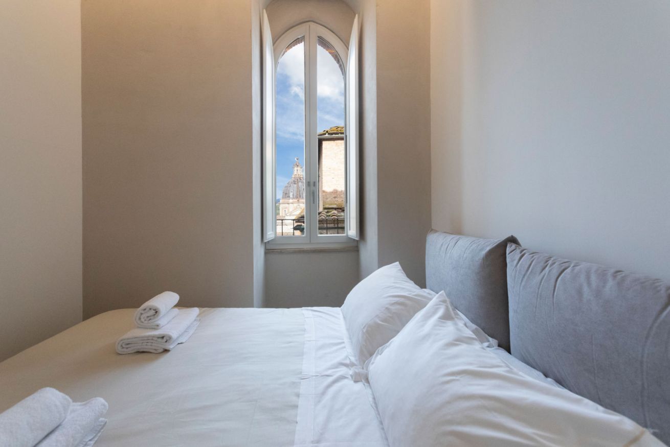 A hotel room with a bed, a window with a view of a building, and a small window above the bed.