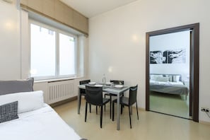 A bedroom with a bed, a window, a dining table with chairs, and a view of a balcony.