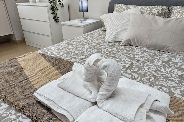 A neatly made bed with patterned bedding, a white bedside table with a lamp, and a small plant.