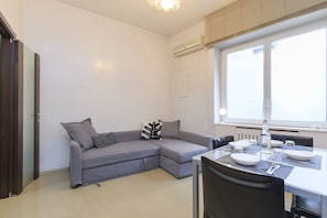 A modern living room with a grey sectional sofa, a dining table with chairs, and a large window providing natural light.