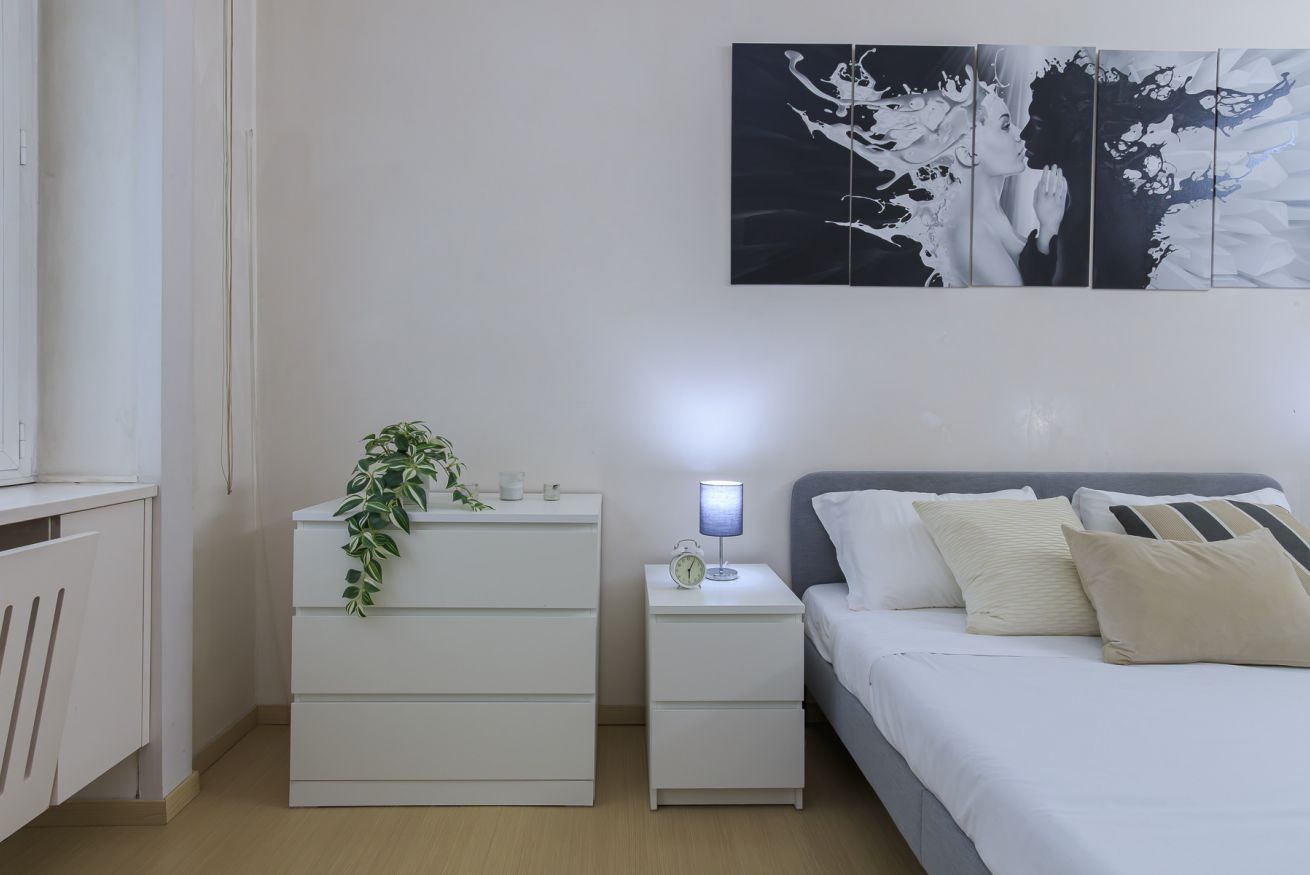 Une chambre à coucher avec un lit, une table de chevet avec une lampe, une commode agrémentée d’une plante et une œuvre d’art murale.