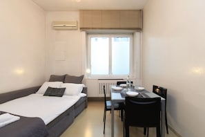 A small, well-lit room with a bed, a dining table with chairs, a window with blinds, and a radiator.