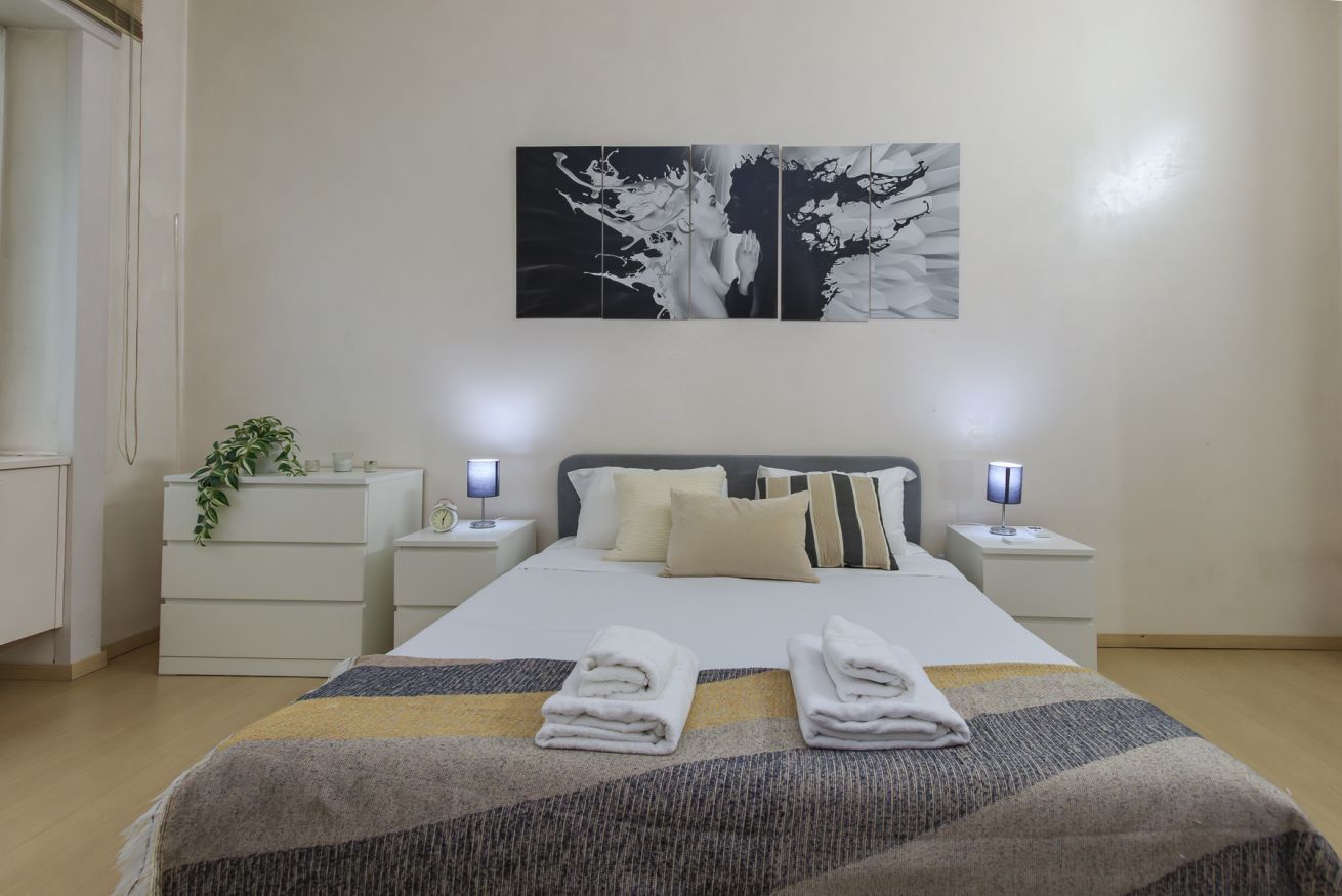 A hotel room with a bed, two bedside tables, a nightstand, a wall-mounted artwork, and a potted plant.