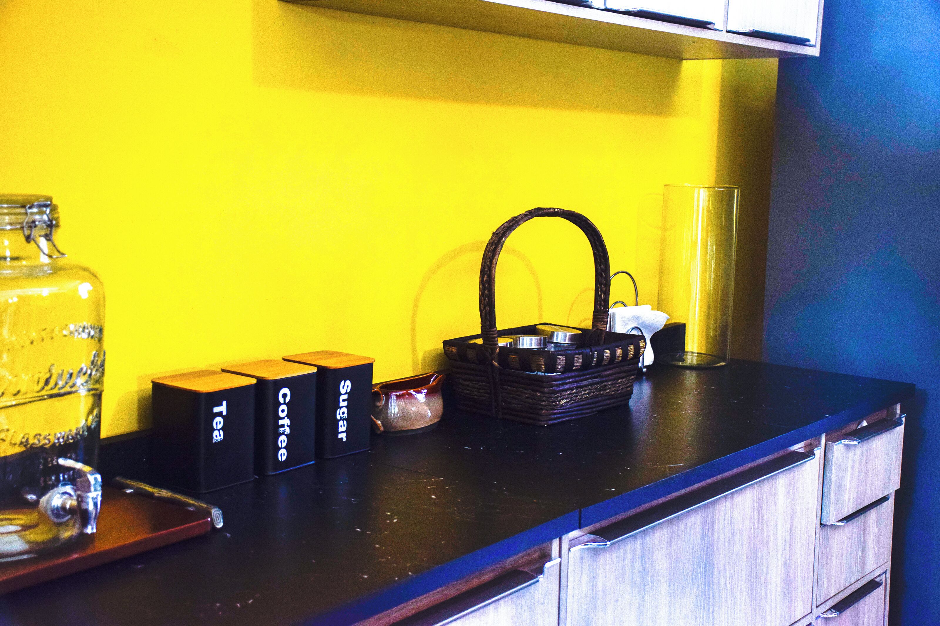 Una encimera de cocina con una pared amarilla, un frasco de vidrio, una cesta y tres cajas etiquetadas como “Tea”, “Coffee” y “SUGAR”.