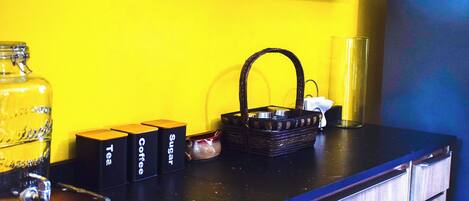 Una encimera de cocina con una pared amarilla, un frasco de vidrio, una cesta y tres cajas etiquetadas como “Tea”, “Coffee” y “SUGAR”.