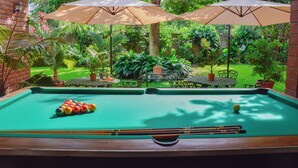 A pool table with balls, surrounded by two umbrellas and a lush garden.