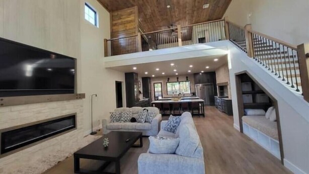 Ein geräumiger Wohnbereich mit einem großen Flachbildfernseher, einer Couch und einem Couchtisch. Eine Treppe führt zu einer oberen Ebene mit einem Balkon.