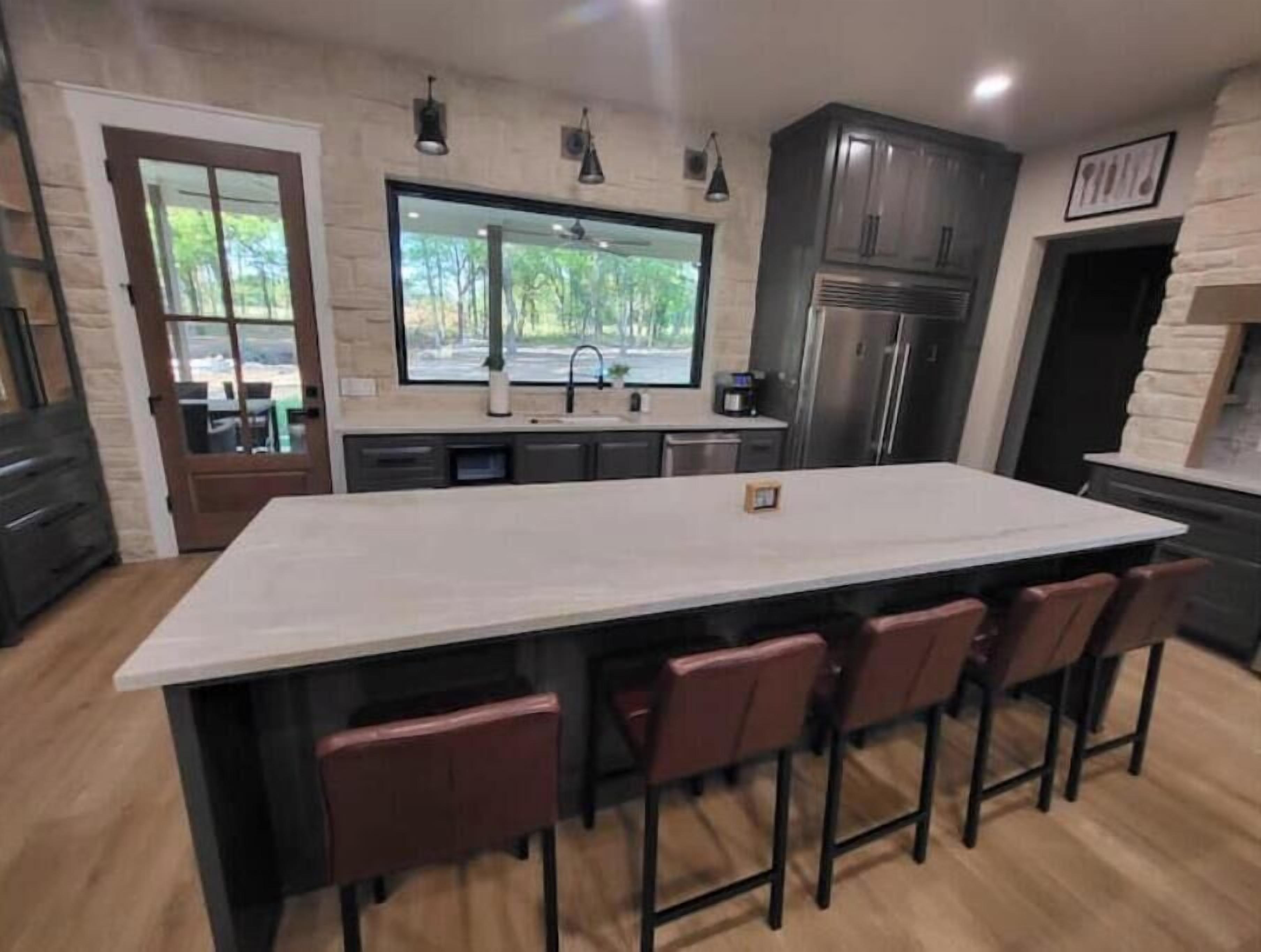 A modern kitchen with a large island, bar stools, and a view of greenery outside.