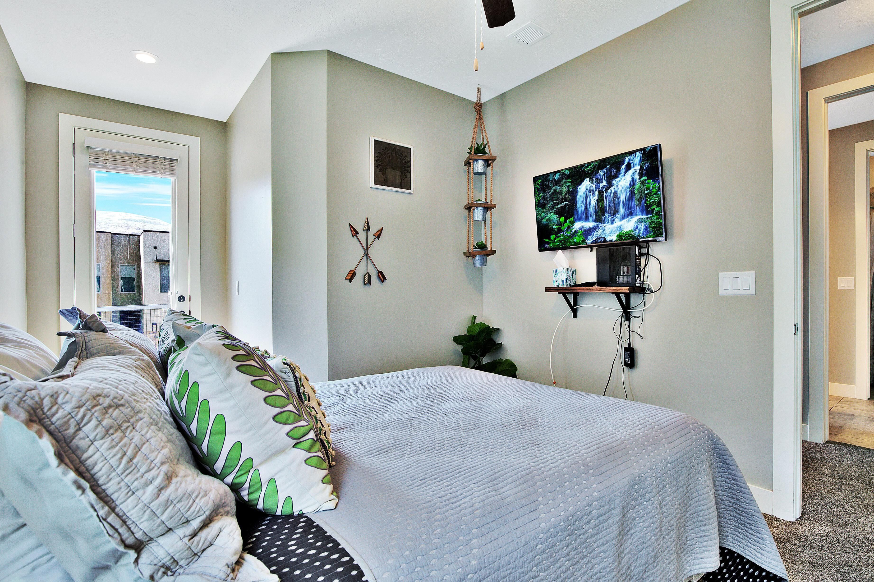 A bedroom with a bed, a TV mounted on the wall, and a window with blinds.