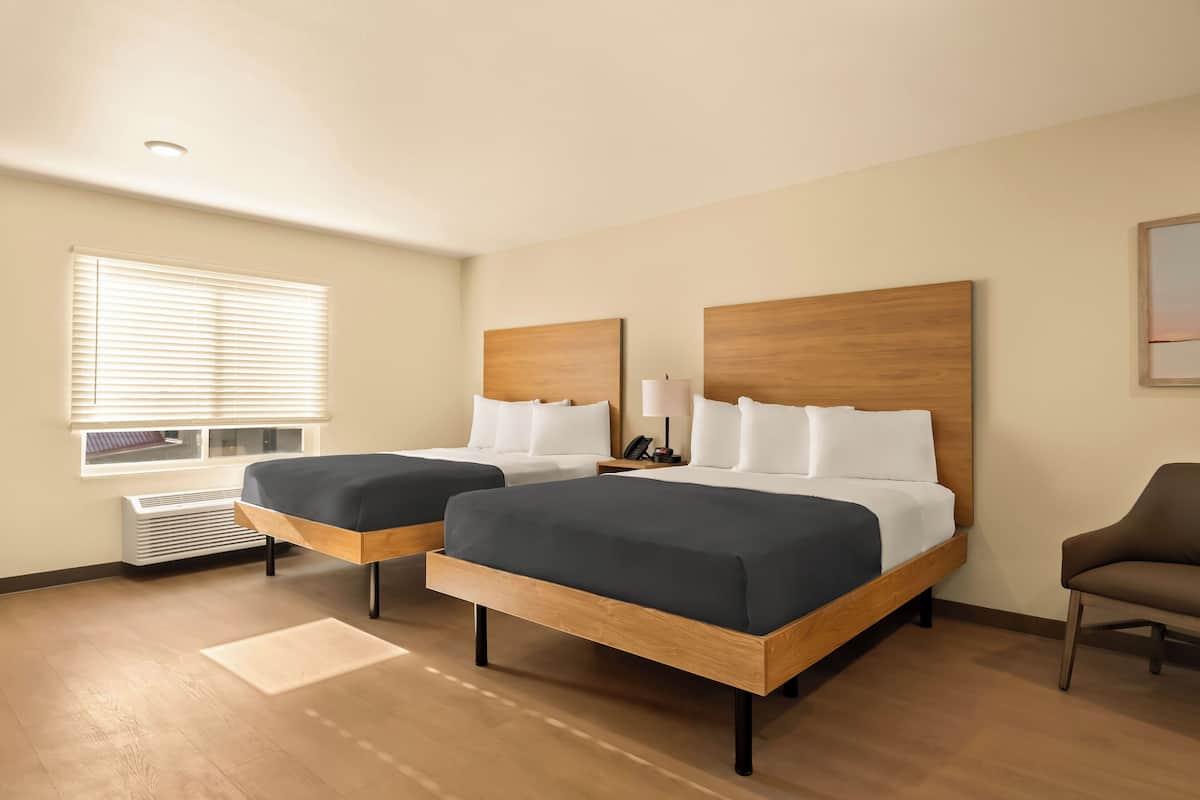 Une chambre d’hôtel avec deux lits, une fenêtre avec des stores, une chaise et une table de chevet avec une lampe.