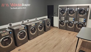 Een wasserette met meerdere wasmachines en drogers, een bord met de tekst 'WASHAWAY', en een houten vloer.