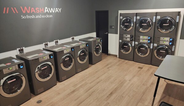 Een wasserette met meerdere wasmachines en drogers, een bord met de tekst 'WASHAWAY', en een houten vloer.