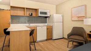 Una cocina compacta equipada con microondas, refrigerador y lavavajillas. Cuenta con una encimera de madera, una silla y una pequeña mesa con lámpara.