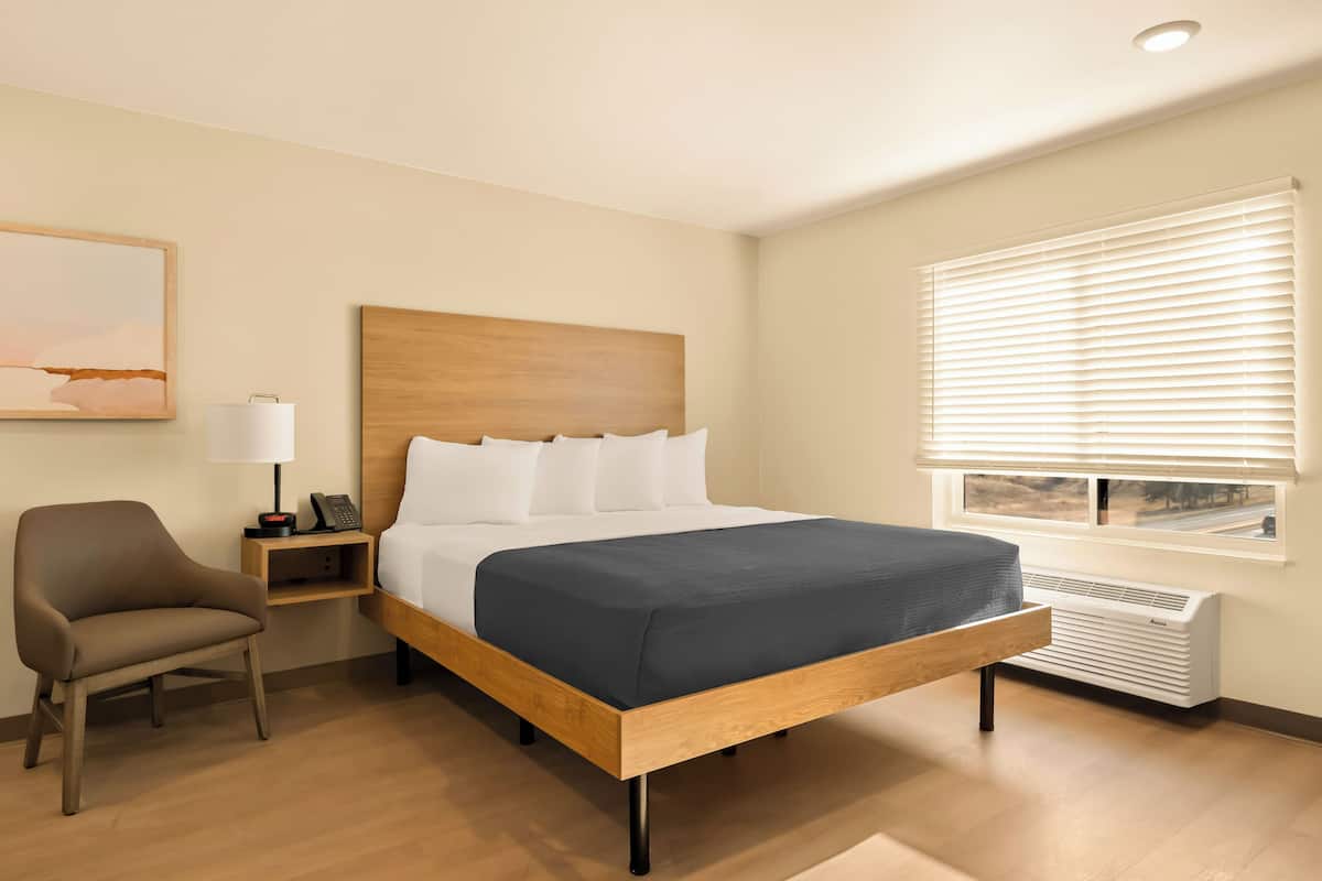 Une chambre d’hôtel avec un lit en bois, des oreillers blancs, une table de chevet avec une lampe et une fenêtre avec des stores.