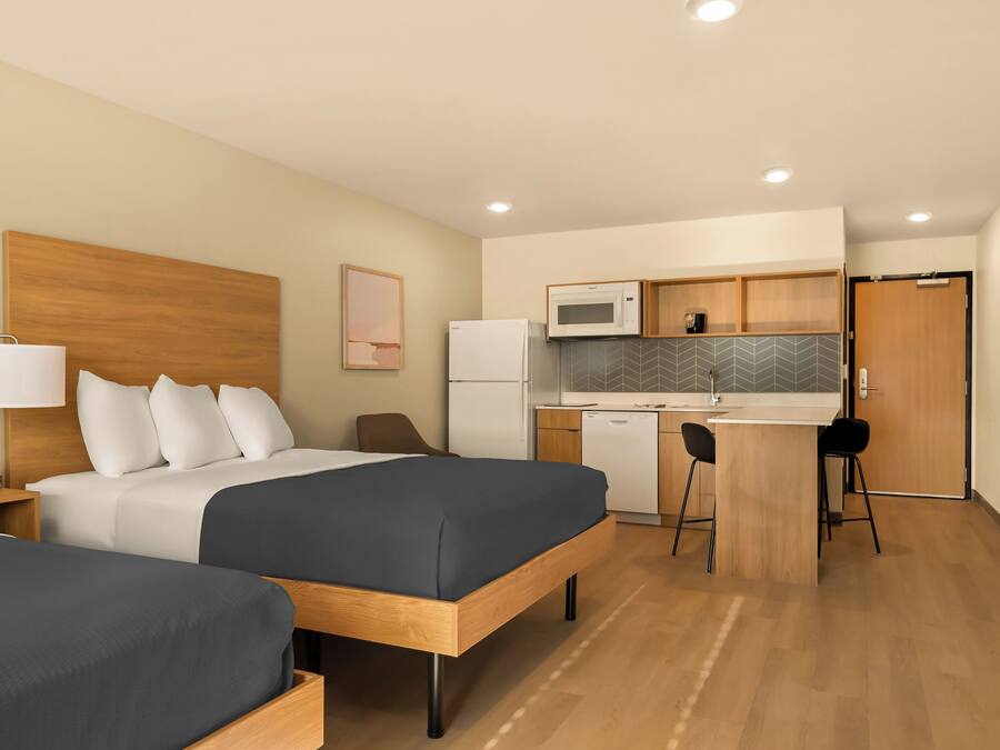 A hotel room with a bed, bedside table, lamp, and a kitchenette with a microwave and refrigerator.