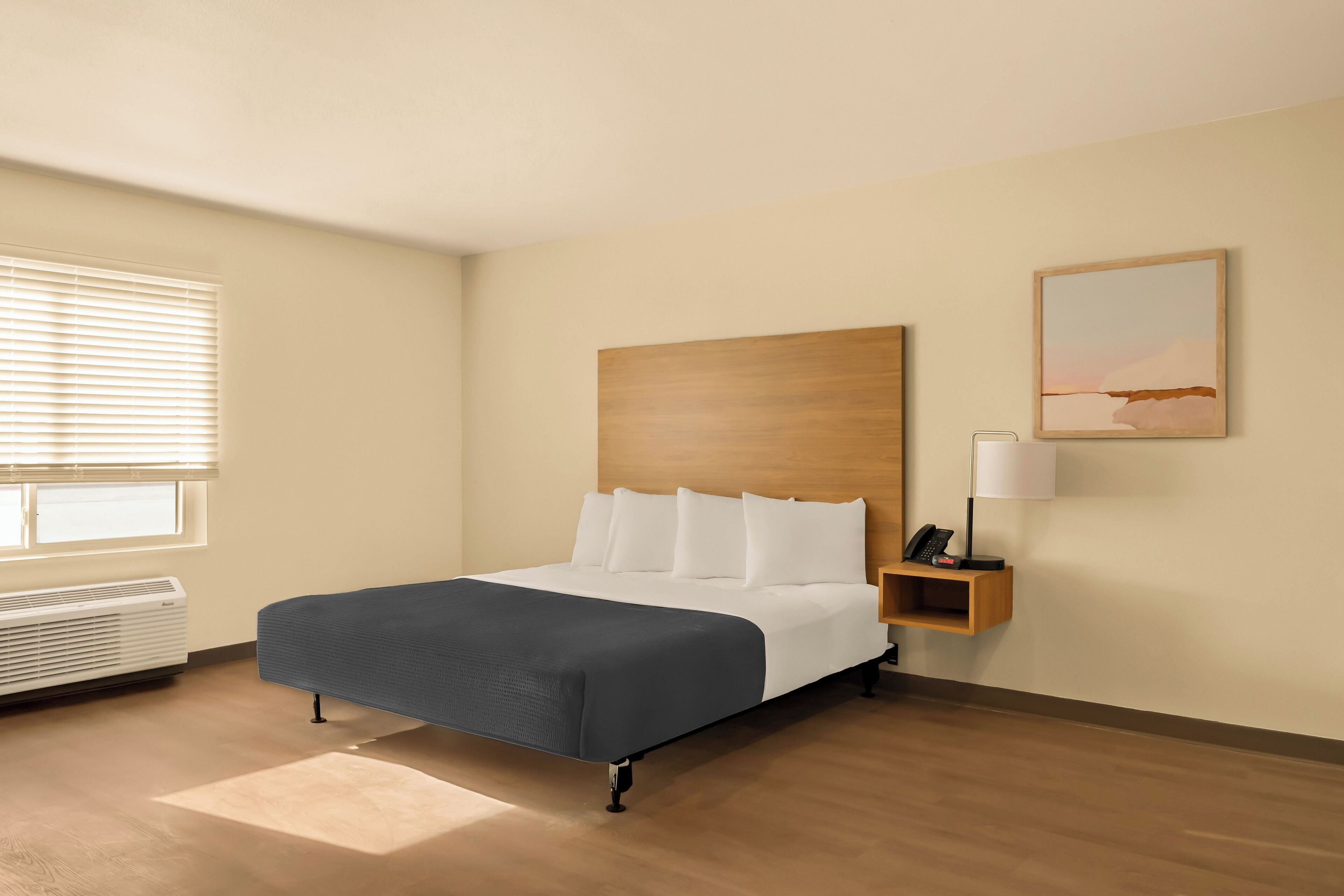 A hotel room with a large bed, a bedside table with a lamp, a painting on the wall, and a window with blinds.