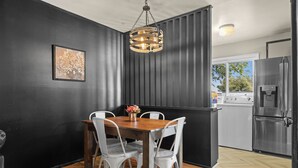 A dining area with a wooden table and white chairs, a hanging light fixture, and a kitchen visible through an open door.