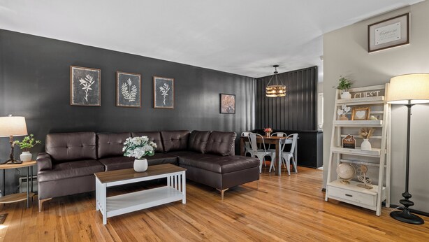 A modern living room with a dark leather sectional sofa, a white coffee table, and a wooden floor. There are framed botanical prints on the wall, a chandelier, and a lamp on a side table.