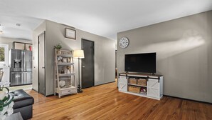 A modern living room with a flat-screen TV on a white cabinet, wooden flooring, a kitchen in the background, and a door leading to another room.
