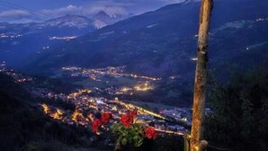 Un banco de piedra con una planta en maceta y luces colgantes, con vistas a un valle iluminado y montañas al anochecer.
