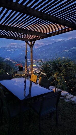 A table with chairs under a pergola with a view of a town and mountains.