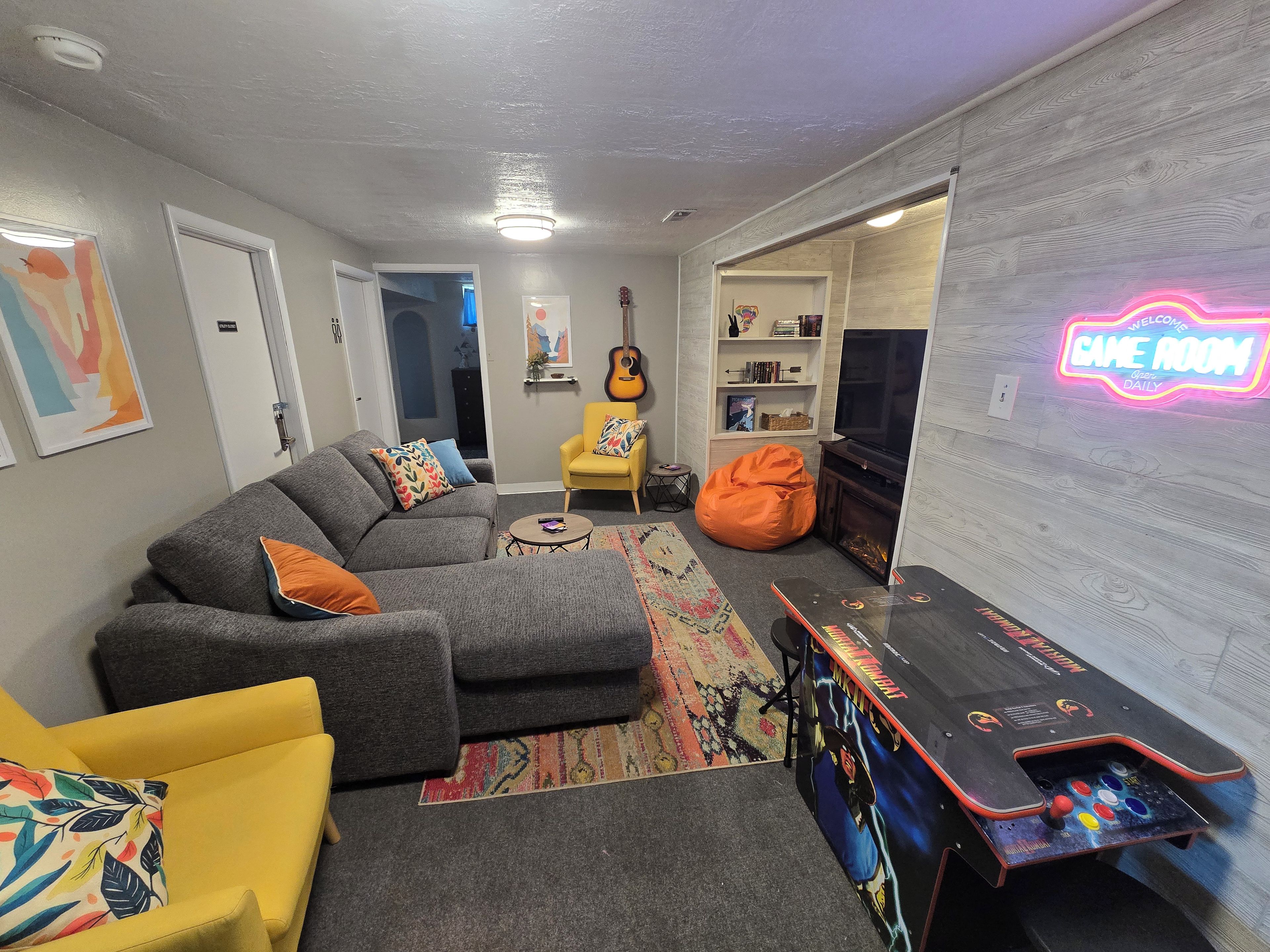 A living room with a sofa, a yellow armchair, a bean bag, a TV, and a neon sign.