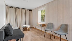 Ein Zimmer mit Holzboden, einem Schlagzeug, einem grauen Sofa, zwei grauen Sesseln und einem Fenster mit Blick auf Grünflächen.