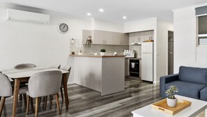 A modern kitchen with a central island, a dining area with a table and chairs, and a living room with a sofa and a plant.