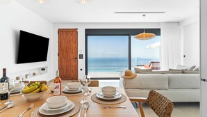 Un espace de repas moderne avec une table en bois dressée pour le dîner, un téléviseur fixé au mur et une vue sur l’océan à travers de grandes fenêtres.