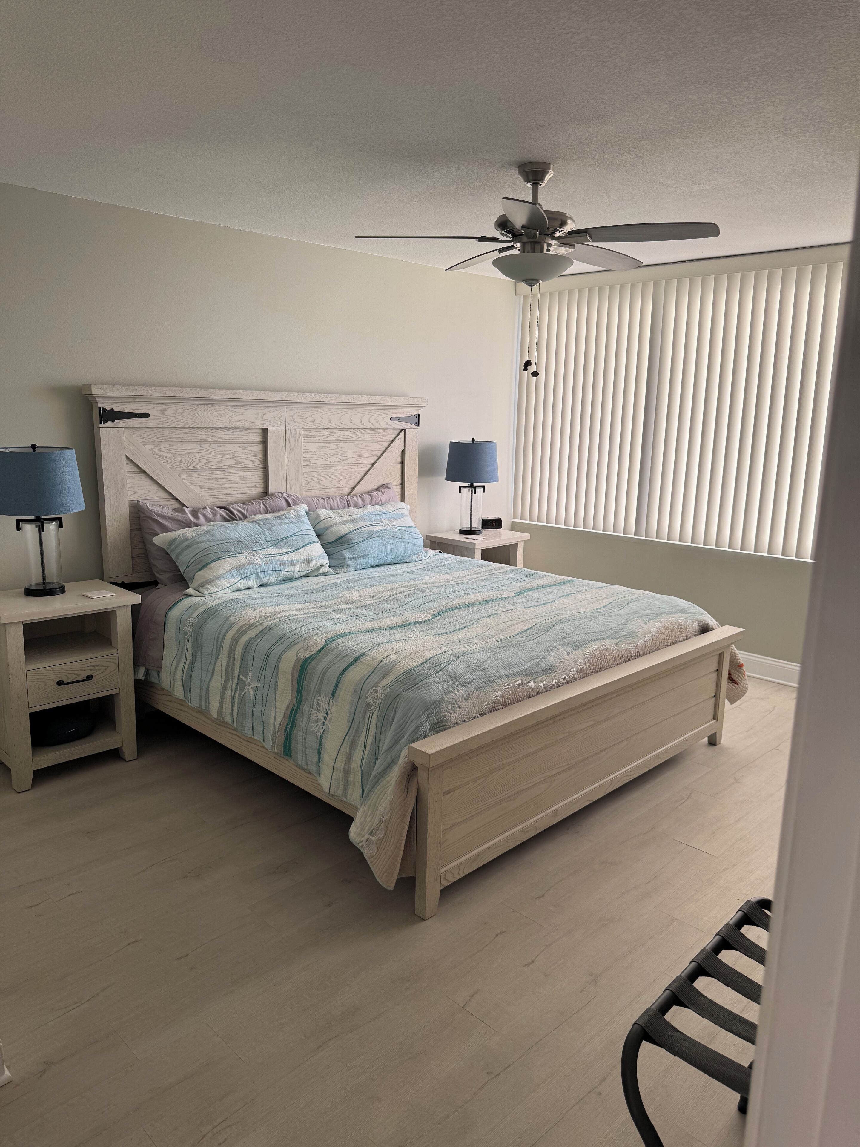 Une chambre à coucher comprenant un lit, deux tables de chevet, un ventilateur de plafond et une fenêtre avec des stores.