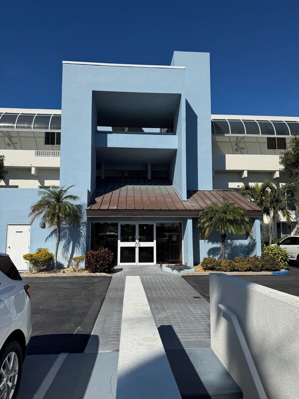 Un edificio moderno con fachada azul, una entrada central con puertas de vidrio y un área de estacionamiento con un coche blanco.