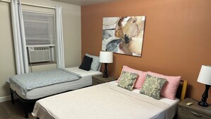 Une chambre d’hôtel avec deux lits, une fenêtre avec des stores, un tableau au mur et une table de chevet avec une lampe.