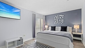 Un dormitorio moderno con una cama grande, un televisor montado en la pared, una mesita de consola y vista al océano a través de la ventana.