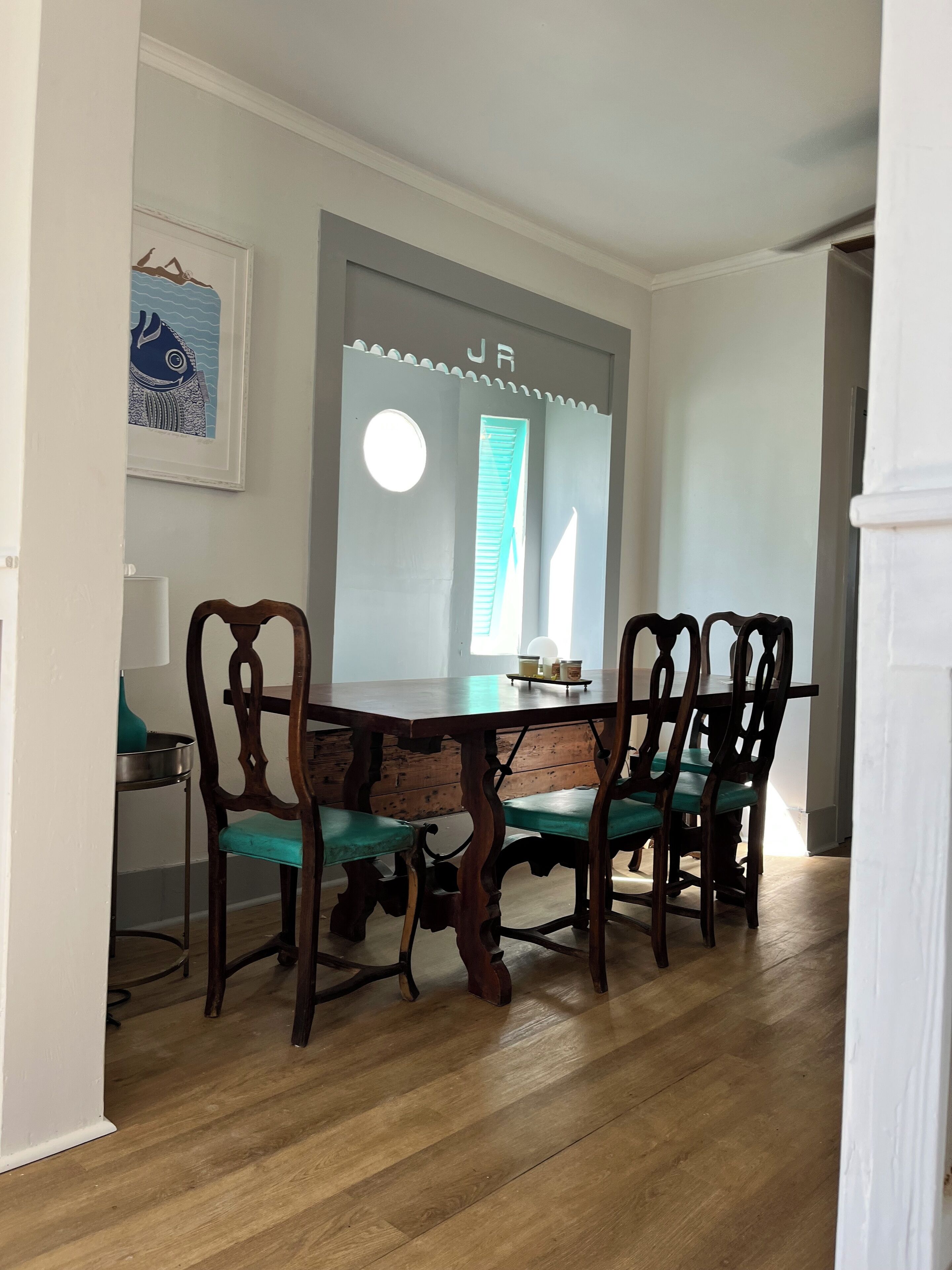 Un comedor con una mesa y sillas de madera, un cuadro en la pared y una ventana con persianas.