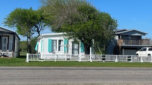 Una casa móvil de un piso con fachada blanca y contraventanas verdes, rodeada por una cerca de piquetes blancos, con un árbol en primer plano.