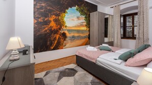 A bedroom with a large mural of a sunset over a rocky coastline, a bed with pink bedding, and a bedside table with a lamp.