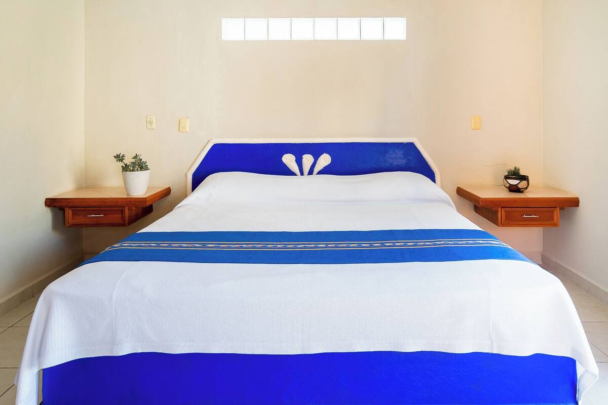 Una cama bien tendida con colcha azul y blanca, dos mesitas de noche con plantas en macetas y una ventana sobre la cama.