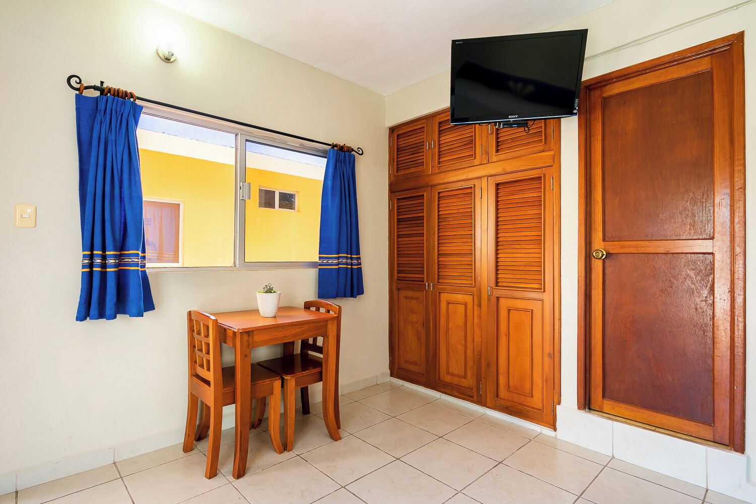 A room with a wooden desk, a TV mounted above a wardrobe, and a window with blue curtains.